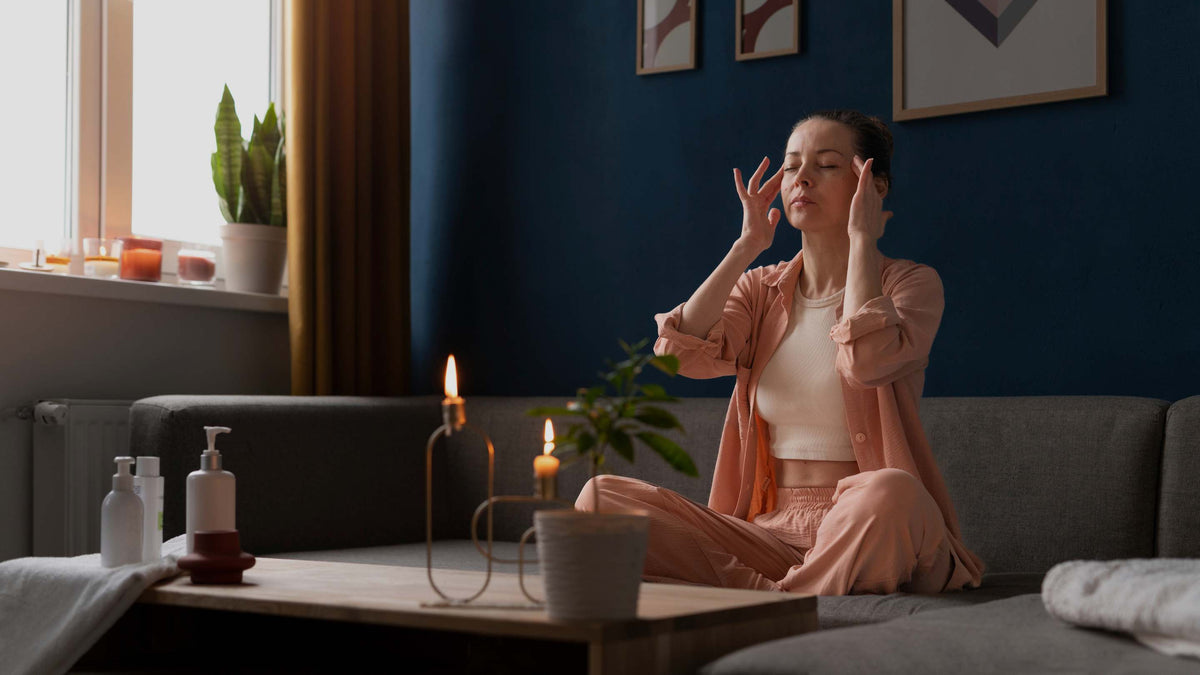A woman in a pink robe sits cross-legged on a gray sofa in a dimly lit room, holding her face with both hands in a serene, meditative pose. A wooden coffee table in the foreground holds lit candles and small bottles, with a dark blue wall and framed art behind her.