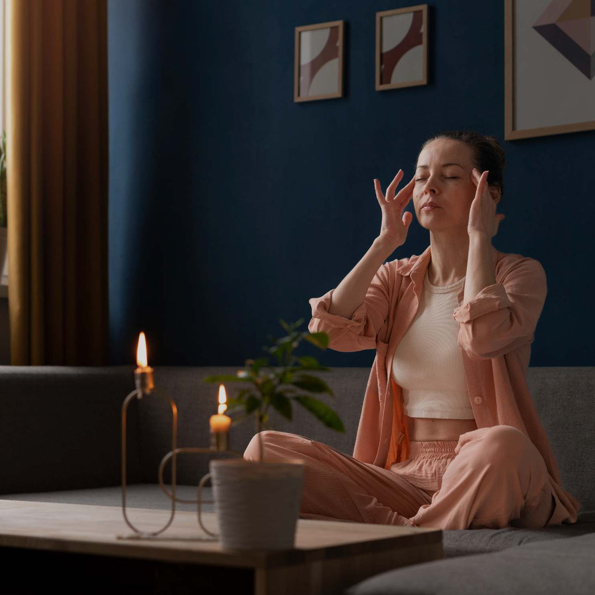 A woman sits cross-legged on a gray sofa in a dimly lit living room, holding her temples with both hands. She is wearing a light pink button-up shirt and matching pink pants over a white crop top.