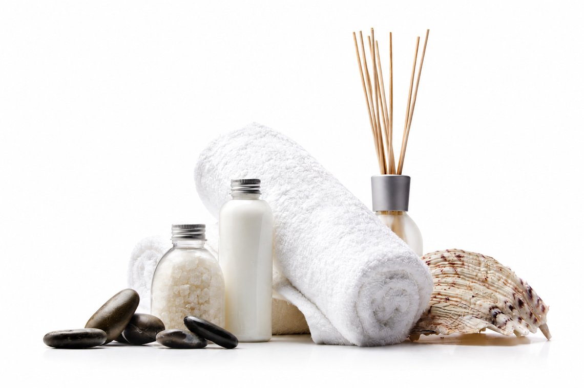 Spa-themed product arrangement on a white background, featuring a white towel, a diffuser with reeds, two small bottles, a seashell, and dark stones.